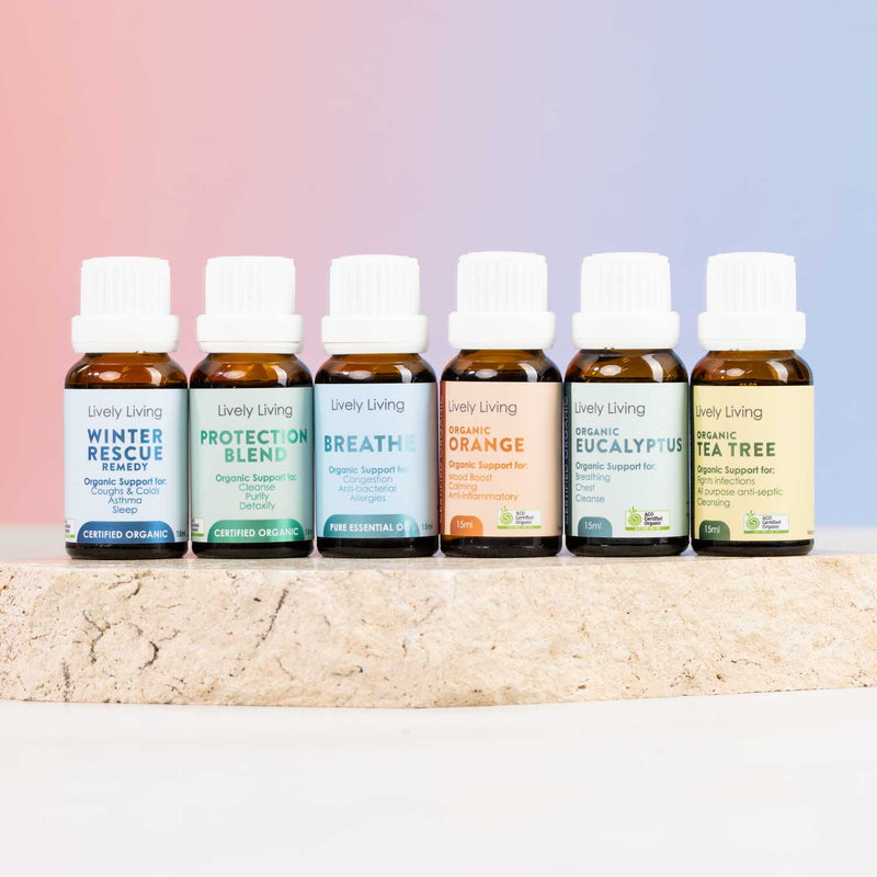 Six small amber essential oil bottles with white caps lined up on a light stone surface against a soft pink-to-blue gradient background. The labels read Lively Living and include Winter Rescue, Protect of Blend, Breath, Organic Orange, Organic Eucalyptus, and Tea Tree.