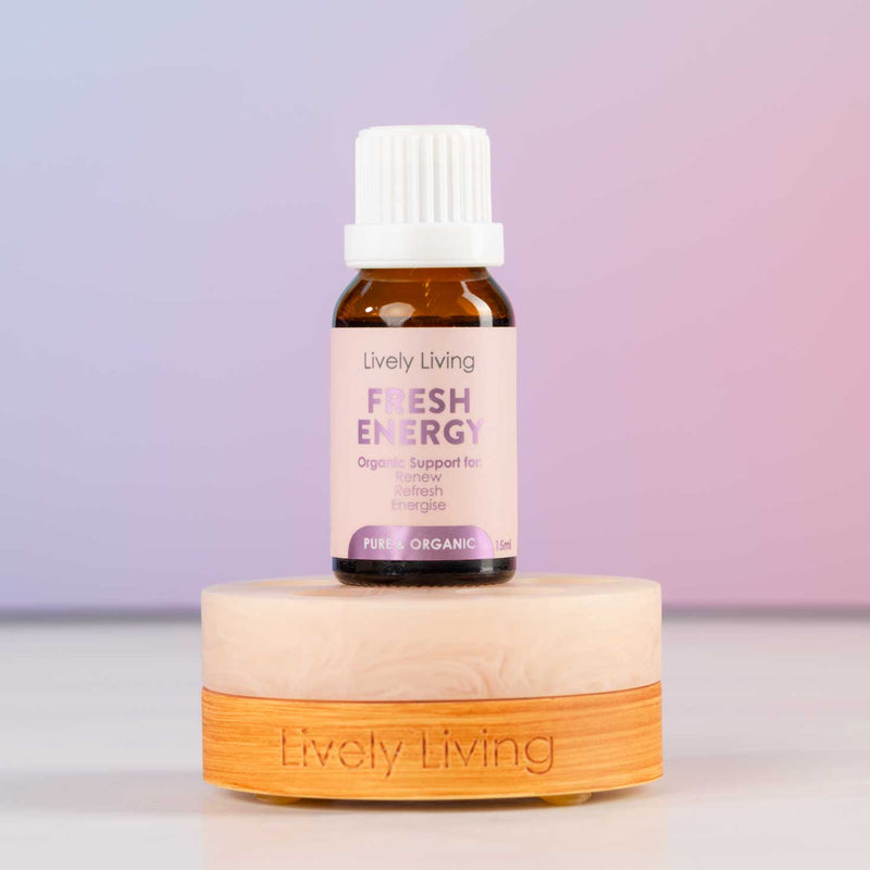 A small amber glass bottle with a white cap and a pink label reading 'Lively Living Fresh Energy' sits on a round light wood base engraved with 'Lively Living.' The background is a soft pastel gradient from lavender to pink.