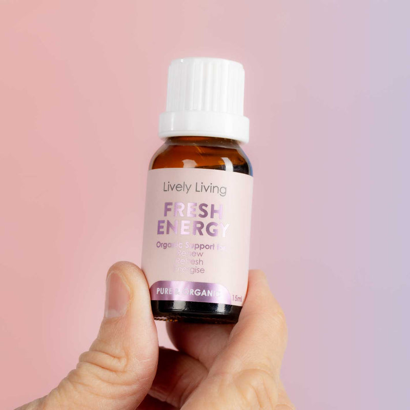 A hand holding a small amber glass bottle of Lively Living Fresh Energy Organic Support Oil against a soft pink and purple gradient background.