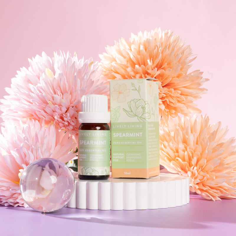 Lively Living "spearmint" essential oil bottle sitting on a white rippled plinth surrounded by pink and orange flowers on a pink background