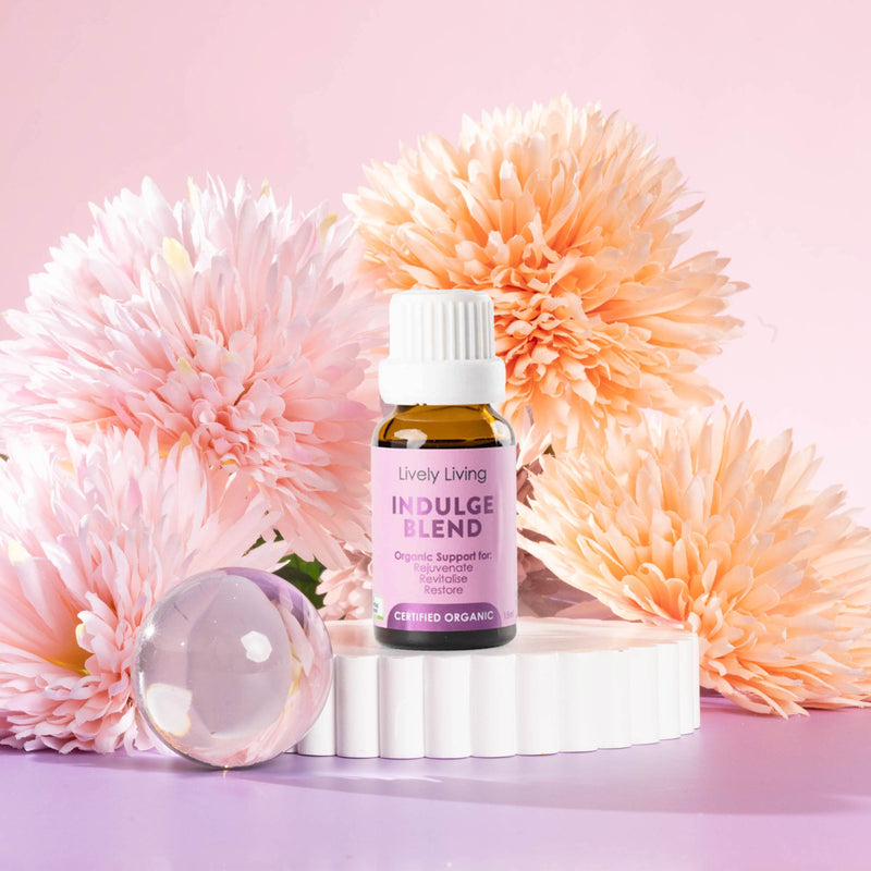 Lively Living "indulge blend" essential oil bottle sitting on a white rippled plinth surrounded by pink and orange flowers on a pink background