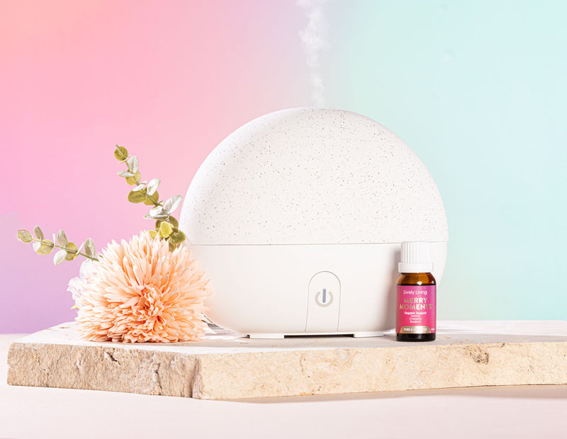 White diffuser with flowers and a bottle on a wooden surface against a pastel gradient background