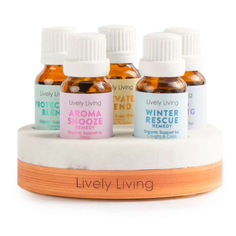 A collection of five small essential oil bottles with white labels, each labeled with different remedy names like Aroma Snooze, Winter Rescue, Elevate, Protection, and Indulge, placed on a wooden stand with the brand name 'Lively Living' at the bottom.