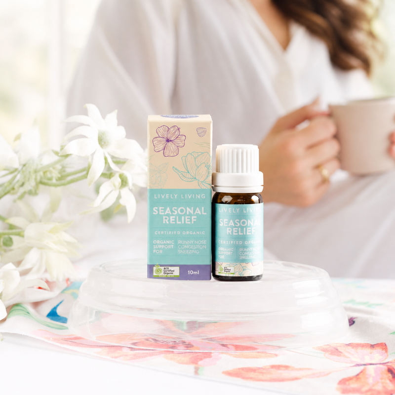 Bottle and box of 'Seasonal Relief' product on a floral blanket with a blurred person holding a mug in the background.