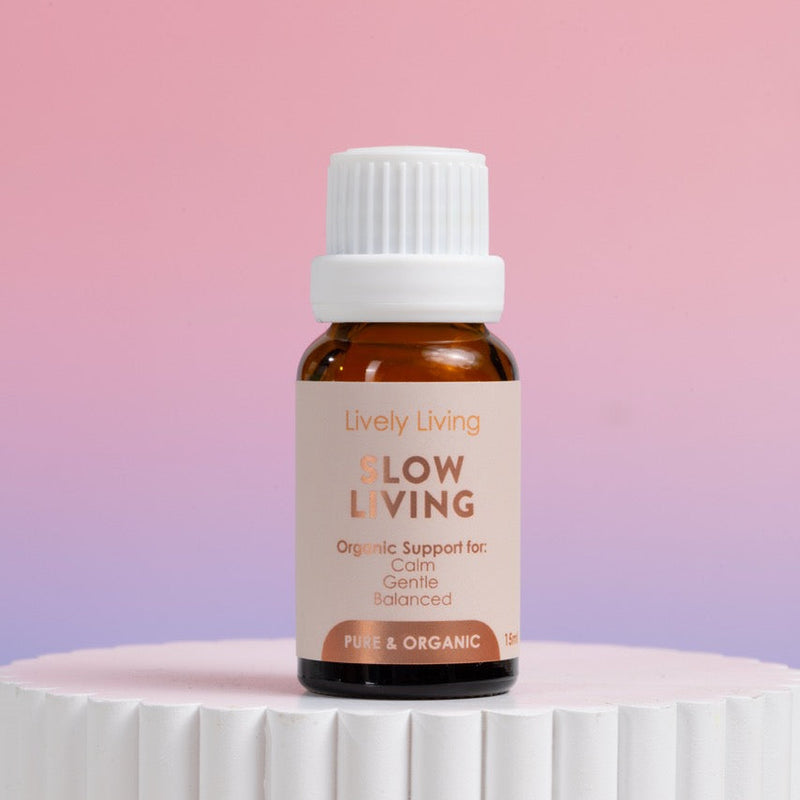 A small amber glass bottle with a white ribbed cap and a beige label reading 'Lively Living Flow Living' stands on a white ribbed pedestal against a soft pink-to-lavender gradient background.