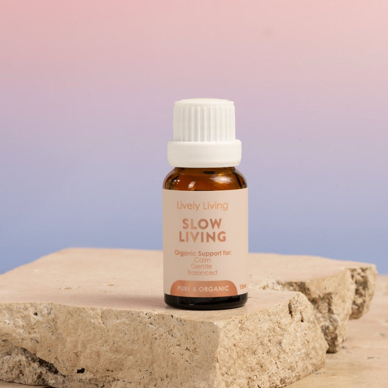An amber glass bottle of 'Slow Living' essential oil with a white cap and beige label, displayed on a textured stone block against a pink and blue gradient background.