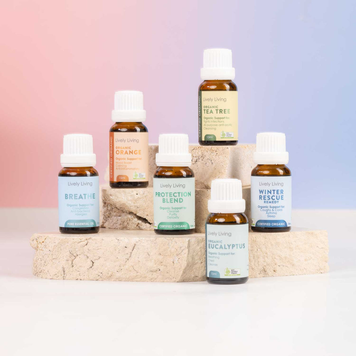Six small amber glass essential oil bottles with white caps from Lively Living are arranged on stacked stone blocks against a soft pink-to-blue gradient background. Visible labels include Breathe, Protection Blend, Winter Rescue, Tea Tree, Eucalyptus, and Orange.