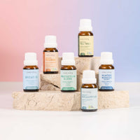 Six small amber glass essential oil bottles with white caps from Lively Living are arranged on stacked stone blocks against a soft pink-to-blue gradient background. Visible labels include Breathe, Protection Blend, Winter Rescue, Tea Tree, Eucalyptus, and Orange.