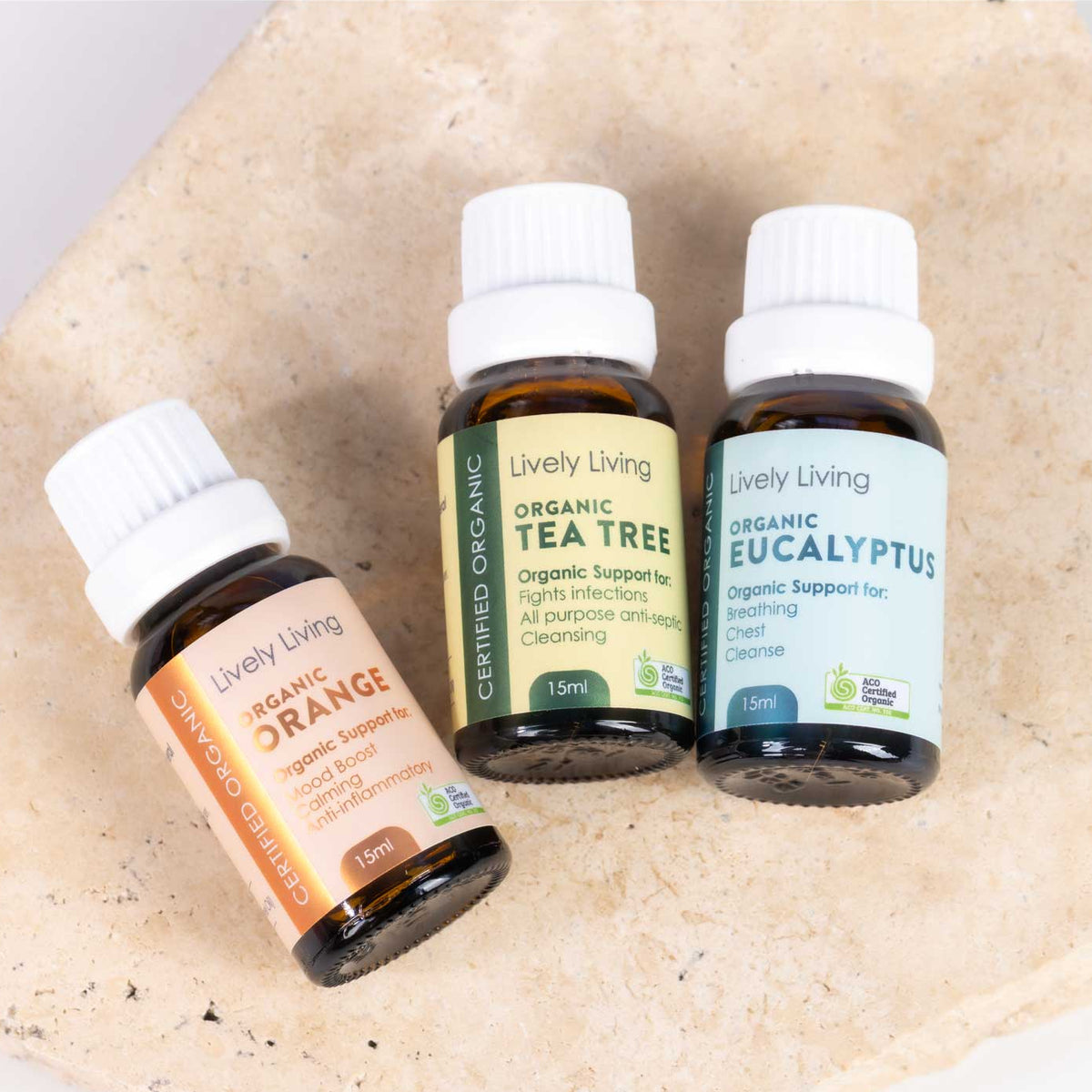 Three small amber glass essential oil bottles with white caps from Lively Living, labeled Organic Orange, Organic Tea Tree, and Organic Eucalyptus, arranged on a light beige stone surface.