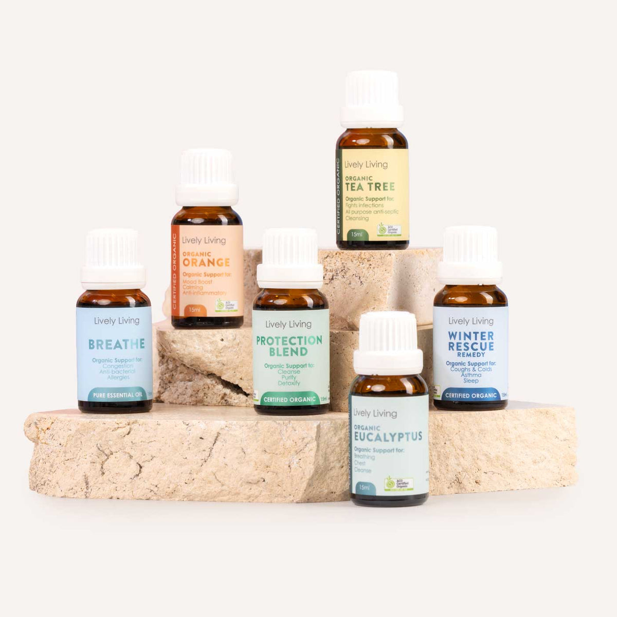 Seven small amber glass essential oil bottles with white caps from the brand Lively Living are arranged on stacked stone blocks against a clean off-white background. The labels show different scents: Breathe, Protection Blend, Tea Tree, Orange, Winter Rescue, Eucalyptus, and Winter Rescue.