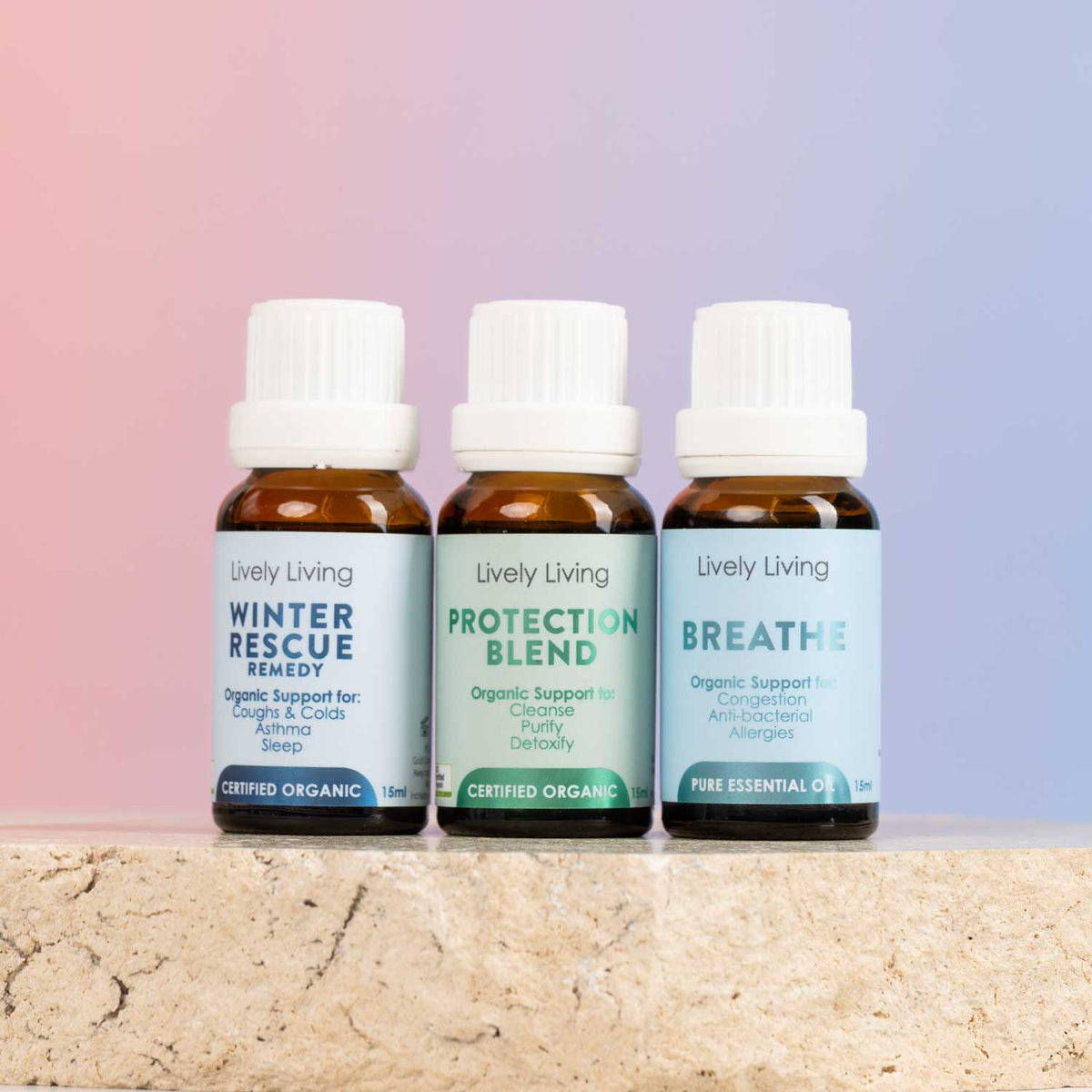 Three amber glass essential oil bottles with white caps from Lively Living, arranged on a stone surface against a soft pink-to-blue gradient background. The labels read Winter Rescue Remedy, Protection Blend, and Breathe.