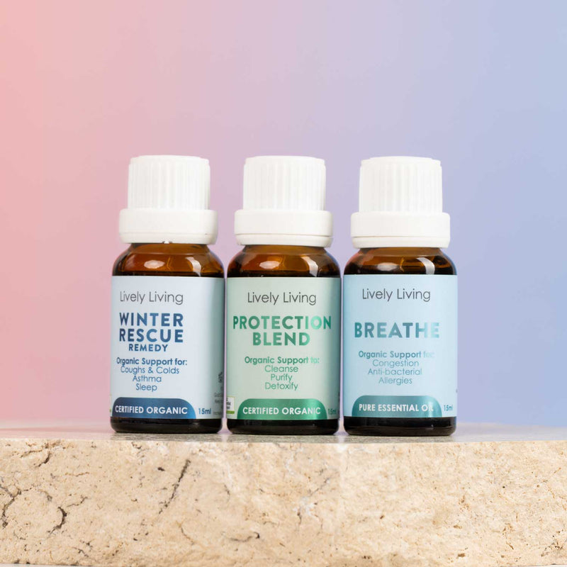 Three amber glass essential oil bottles with white caps from Lively Living, arranged on a stone surface against a soft pink-to-blue gradient background. The labels read Winter Rescue Remedy, Protection Blend, and Breathe.