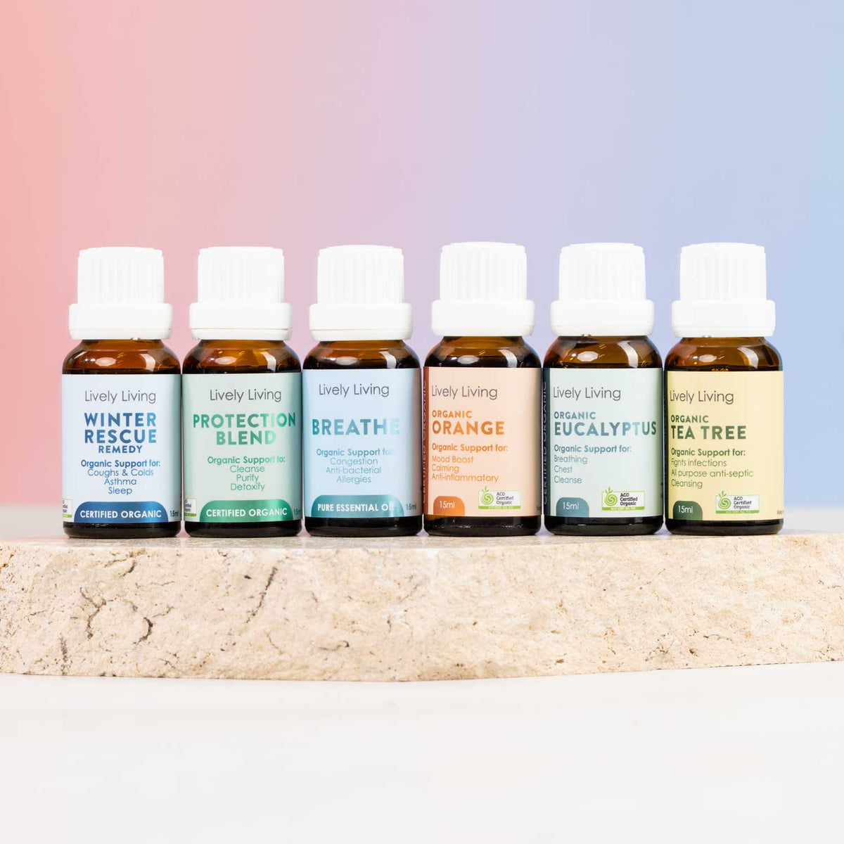 Six small amber essential oil bottles with white caps lined up on a light stone surface against a soft pink-to-blue gradient background. The labels read Lively Living and include Winter Rescue, Protect of Blend, Breath, Organic Orange, Organic Eucalyptus, and Tea Tree.