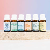 Six small amber essential oil bottles with white caps lined up on a light stone surface against a soft pink-to-blue gradient background. The labels read Lively Living and include Winter Rescue, Protect of Blend, Breath, Organic Orange, Organic Eucalyptus, and Tea Tree.