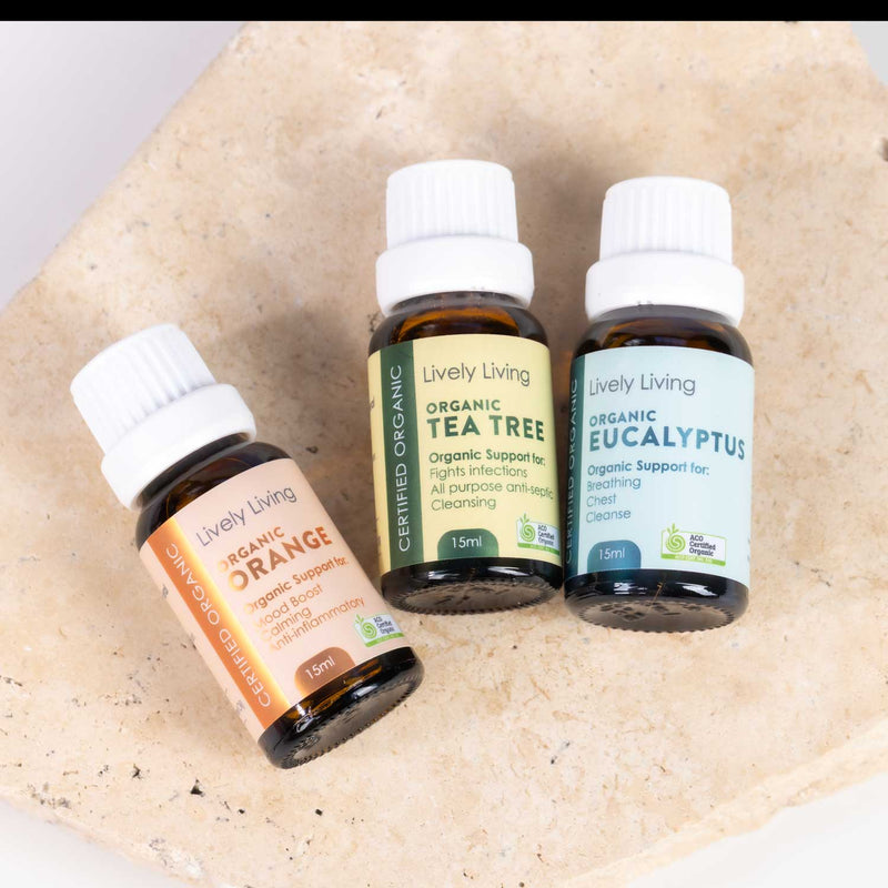 Three small amber glass essential oil bottles with white caps from Lively Living, labeled Organic Orange, Organic Tea Tree, and Organic Eucalyptus, arranged on a light beige stone surface.