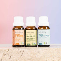 Three small amber glass essential oil bottles with white ribbed caps from Lively Living, arranged on a light stone surface against a soft pink-to-blue gradient background. The bottles are labeled Organic Orange, Organic Tea Tree, and Organic Eucalyptus.