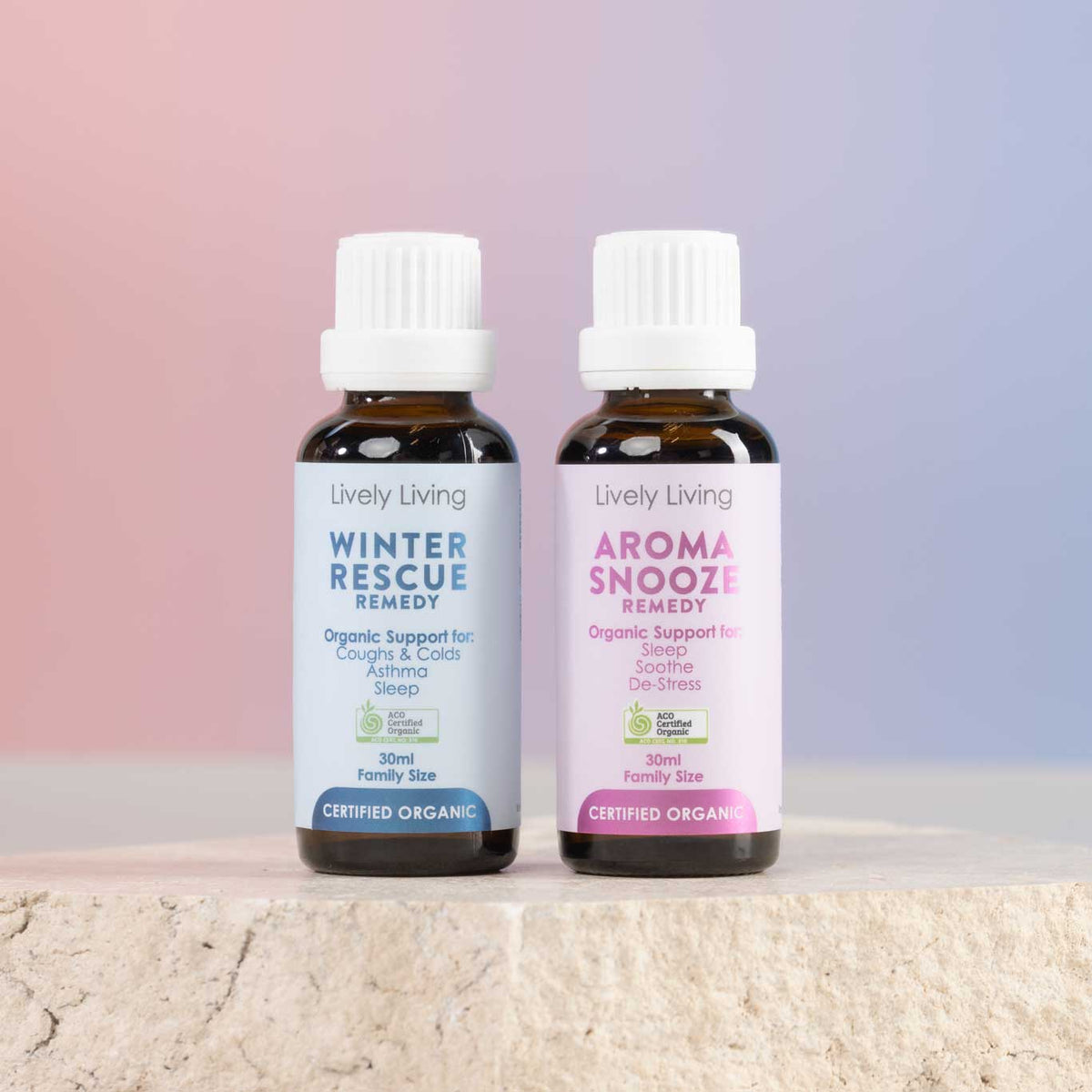 Two small amber bottles of Lively Living essential oil remedies, Winter Rescue Remedy and Aroma Sneeze Remedy, standing side-by-side on a light stone surface against a soft pink and blue gradient background.
