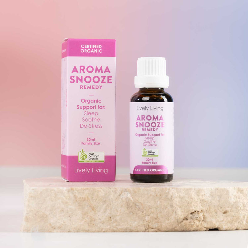 A bottle and box of Lively Living Aromasnooze Remedy organic support blend, displayed on a textured stone slab against a soft pink and blue gradient background.
