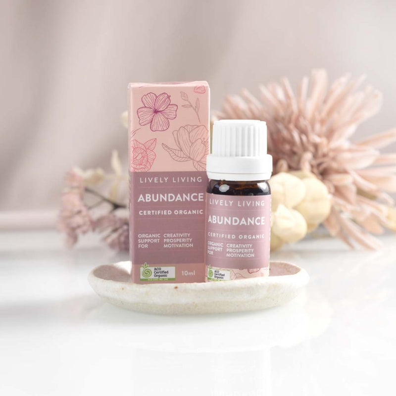 A bottle of the Lively Living "Abundance" essential oil and a label that reads 'Lively Living . Surrounded by a white surface and neutral soft pink florals in the background that are blurred