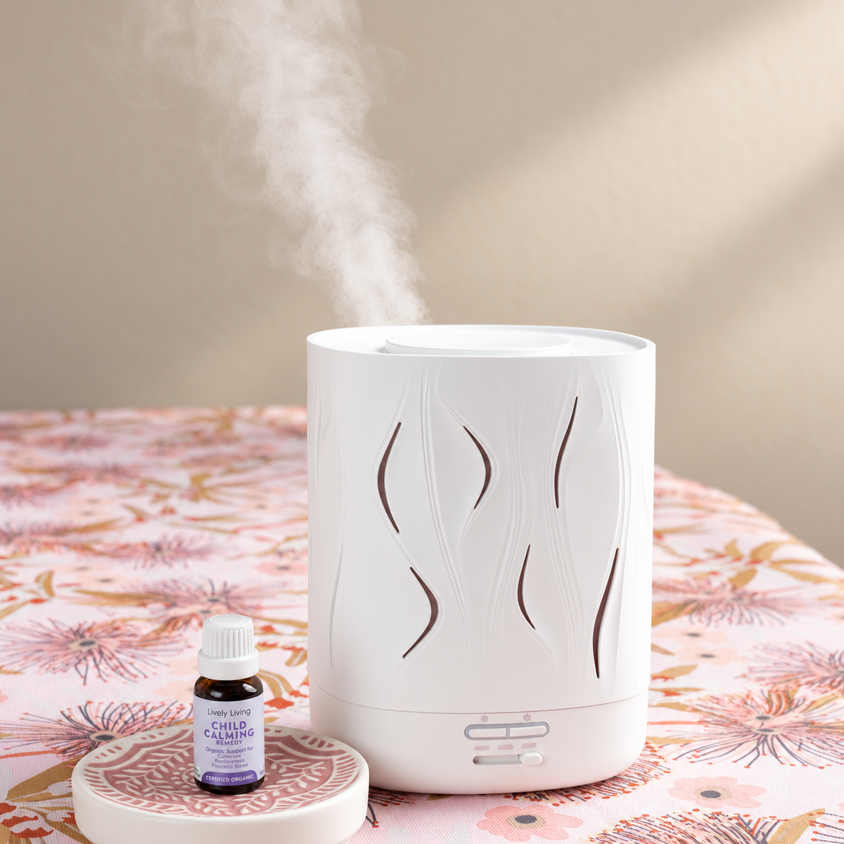 White Aroma-Cloud diffuser releasing mist on a floral tablecloth beside a small bottle of Lively Living oil with a lilac label