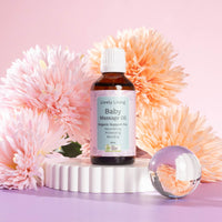 A bottle of the Lively Living Baby Massage Oil with a label that reads 'Lively Living Baby Massage Oil, Organic Support for Nourishing, Protecting, Bonding', with a capacity of 50ml. Surrounded by pink and orange flowers on a rippled white  plinth