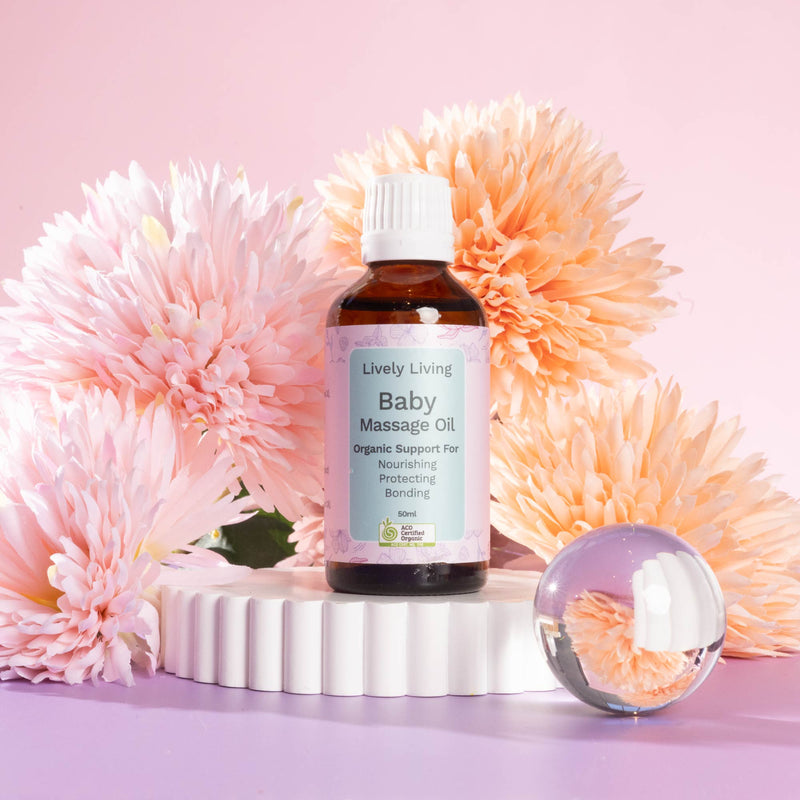 A bottle of the Lively Living Baby Massage Oil with a label that reads 'Lively Living Baby Massage Oil, Organic Support for Nourishing, Protecting, Bonding', with a capacity of 50ml. Surrounded by pink and orange flowers on a rippled white  plinth