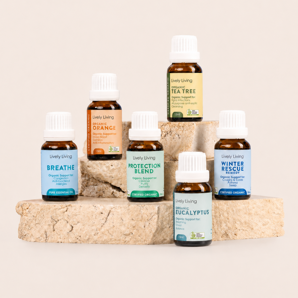 six small amber glass essential oil bottles with white caps from Lively Living are arranged on stacked stone blocks against a warm off-white background. Visible labels include Breathe, Orange, Protection Blend, Winter Rescue, Eucalyptus, Tea Tree, and Winter Blend.