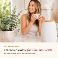 Smiling woman holding a mug at a table beside a white etched ceramic Aroma-Fern diffuser with a bamboo lid releasing mist and a eucalyptus plant, with the text Ceramic calm, for slow moments