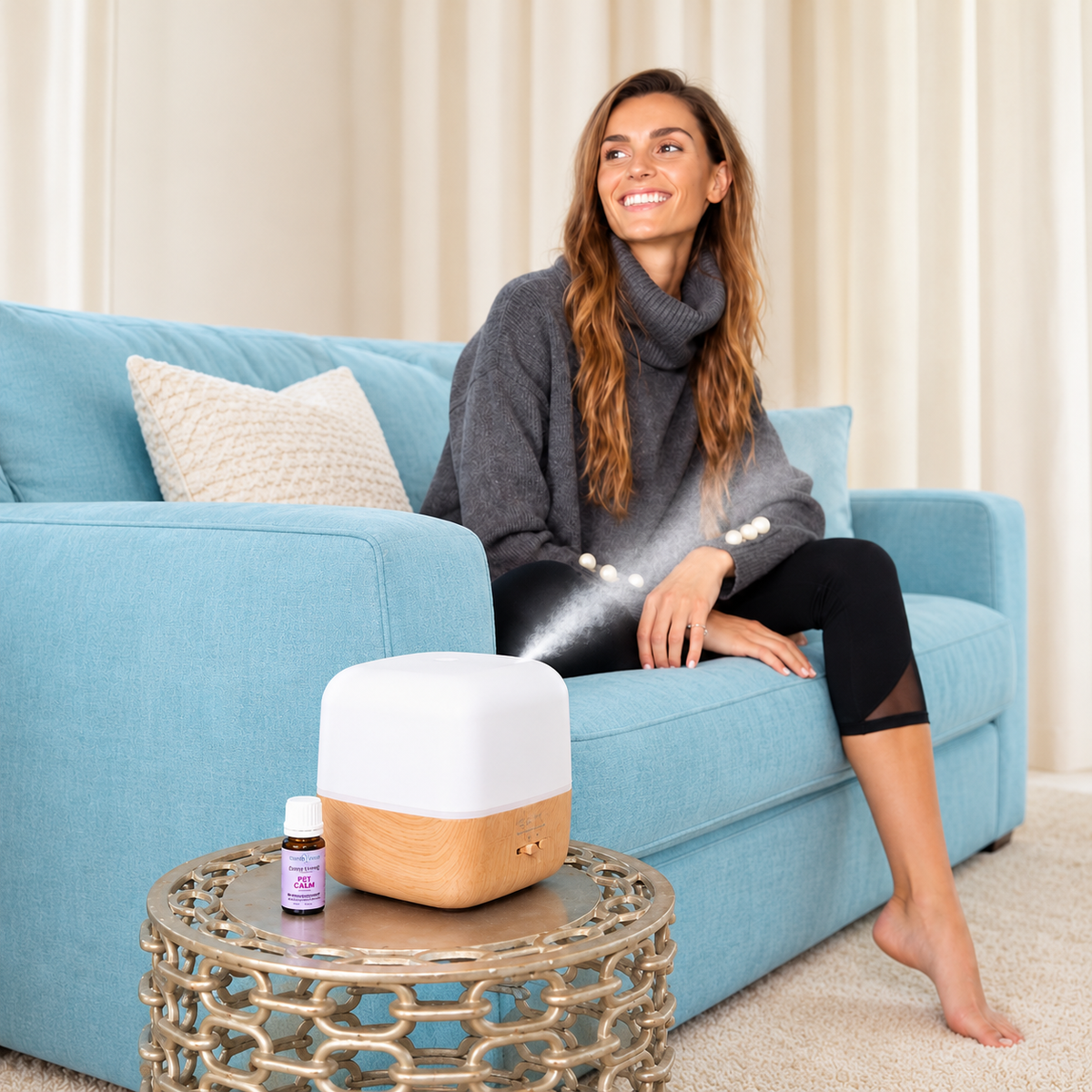 A woman sits on a light blue sofa in a living room setting, smiling while a white and wood diffuser emits a mist from its nozzle. A small essential oil bottle is placed on a decorative gold side table in the foreground.
