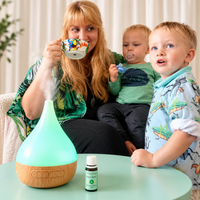 A woman and two children are gathered around a glowing teal diffuser on a mint table, with a small essential oil bottle in front of them. The diffuser is emitting a visible mist.