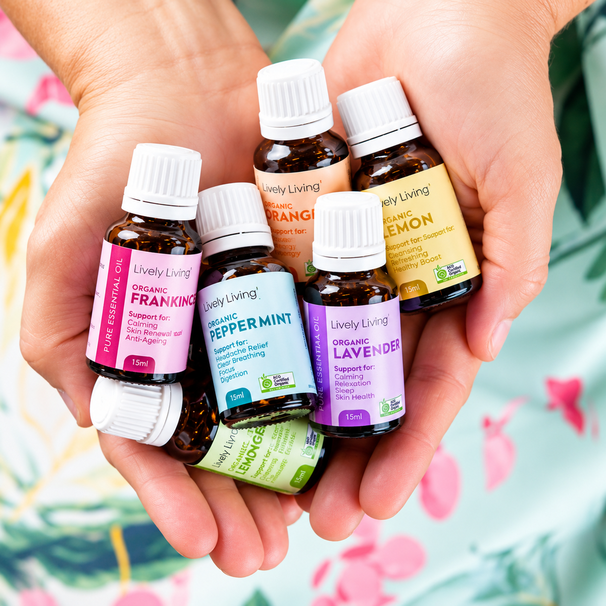 A pair of hands holding six small Lively Living essential oil bottles in both hands against a colorful floral background. The bottles include Organic Frankincense, Organic Peppermint, Organic Lavender, Organic Orange, and Organic Lemon.