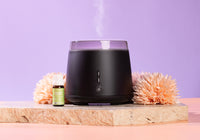 Black essential oil diffuser with a bottle of essential oil and flowers on a light stone surface against a purple background