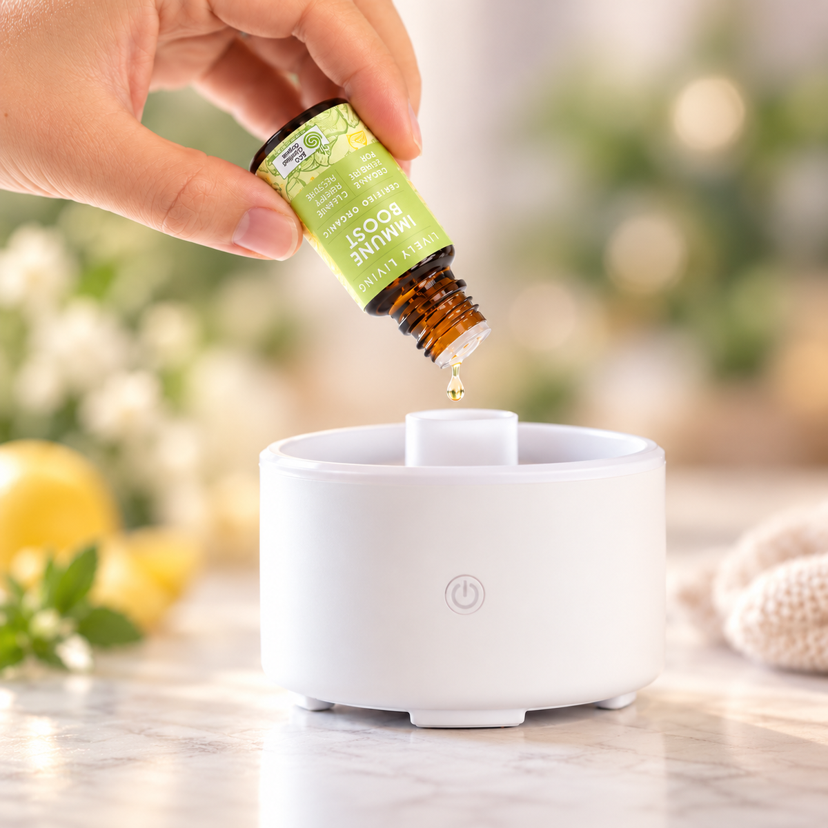 Hand tipping a drop of Lively Living Immune Boost essential oil into a small white diffuser on a marble bench, with lemons and white flowers in the background