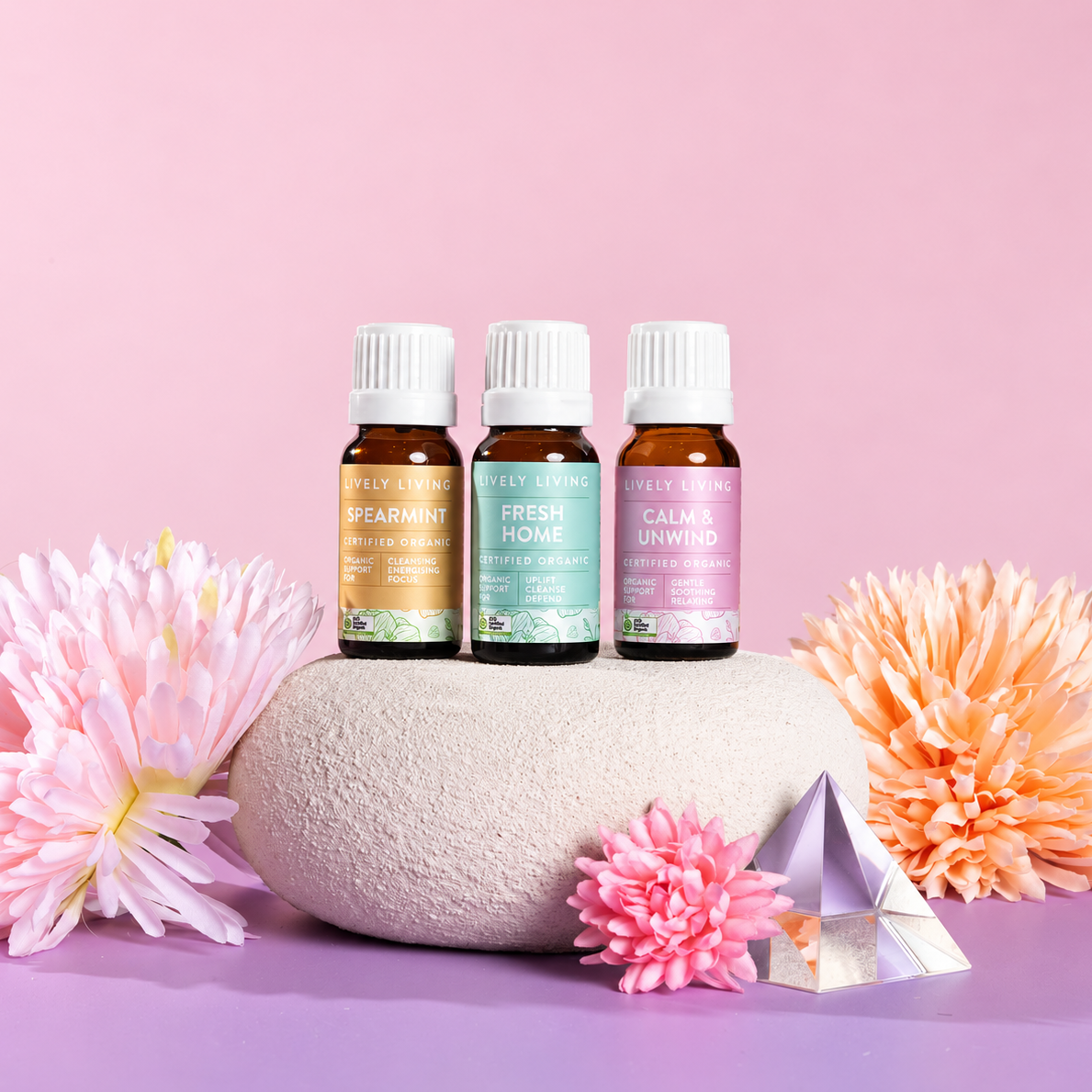 Lively Living Spearmint, Fresh Home and Calm & Unwind oils on a round stone pedestal with pink and peach chrysanthemums and a crystal on a pink and lilac background