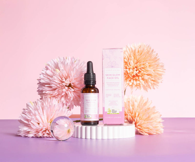 Lively Living "skin glow face oil" essential oil bottle sitting on a white rippled plinth surrounded by pink and orange flowers on a pink background