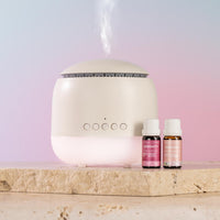 A white diffuser with misting at the top sits on a stone slab next to two small pink and amber essential oil bottles labeled Serenity and Calm.