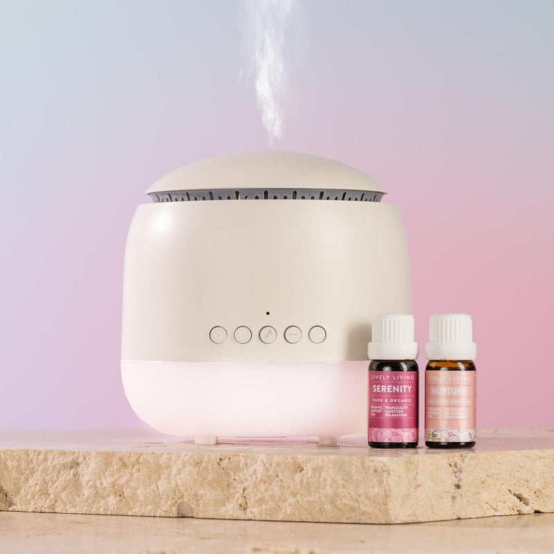 A white diffuser with misting at the top sits on a stone slab next to two small pink and amber essential oil bottles labeled Serenity and Calm.