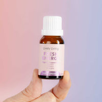 A hand holding a small amber glass bottle of Lively Living Fresh Energy essential oil against a soft pink and purple gradient background.