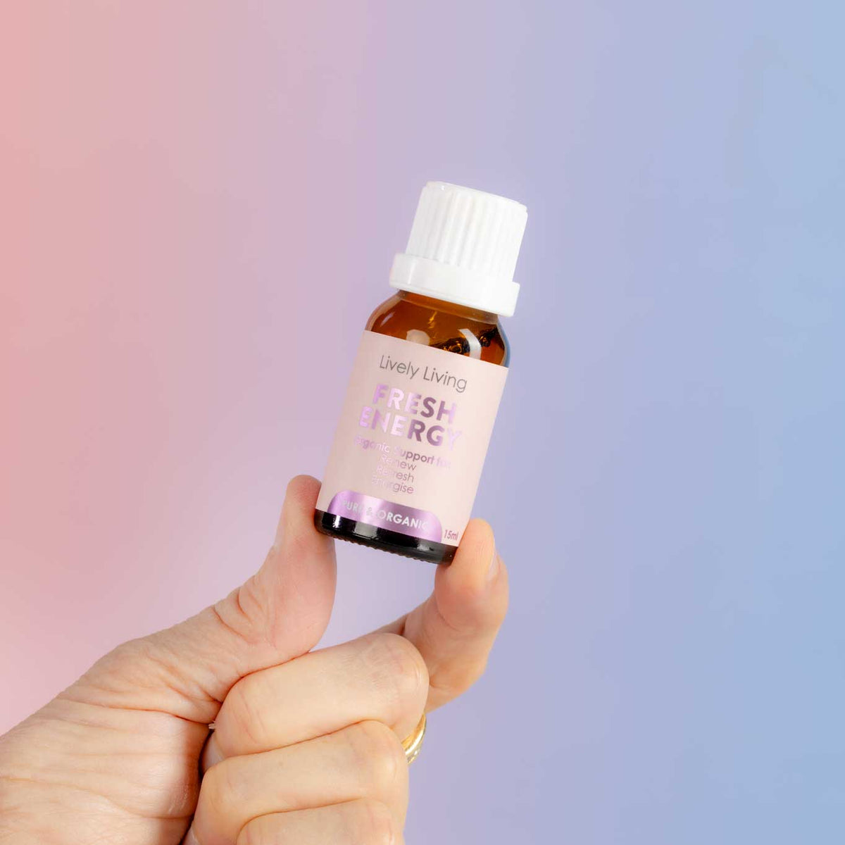 A hand holding a small amber glass bottle of Uvely Living Fresh Energy essential oil against a soft pink-to-blue gradient background.