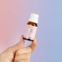 A hand holding a small amber glass bottle of Uvely Living Fresh Energy essential oil against a soft pink-to-blue gradient background.