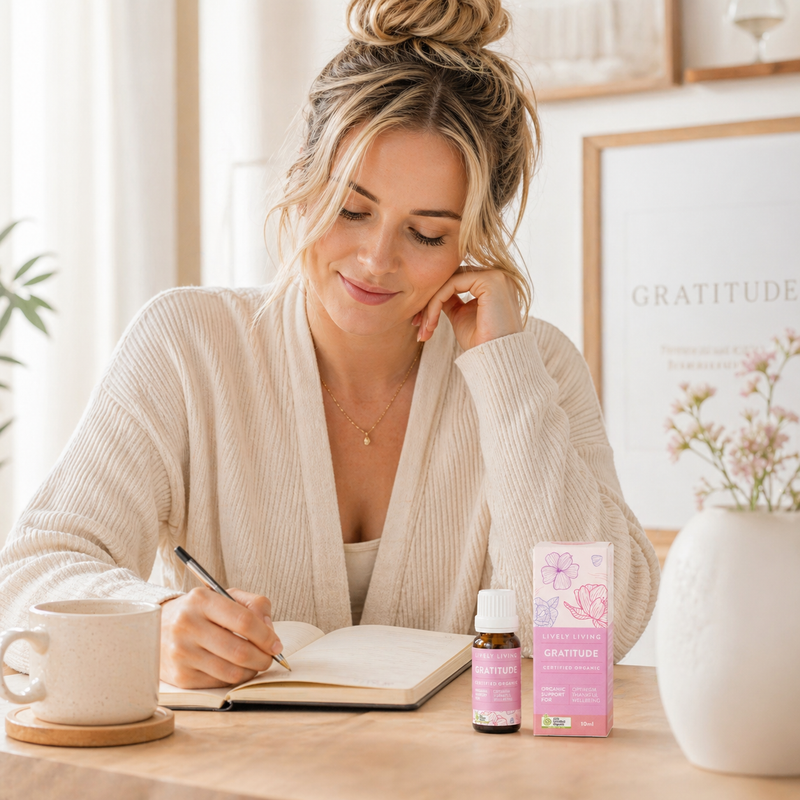 Woman in a robe sitting at a table with a notebook and mug, next to a bottle and box of 'Gratitude' products.