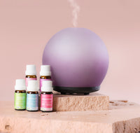 Purple diffuser emitting steam with various essential oil bottles on a wooden surface and pink background