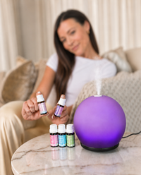 A woman sits on a couch holding two small essential oil bottles beside a glowing purple diffuser and four small bottles on a marble table in a living room setting.