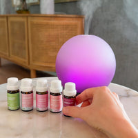 A hand is shown selecting a small essential oil bottle from a row of five bottles lined up on a marble-topped table next to a glowing purple diffuser sphere in a home interior setting.