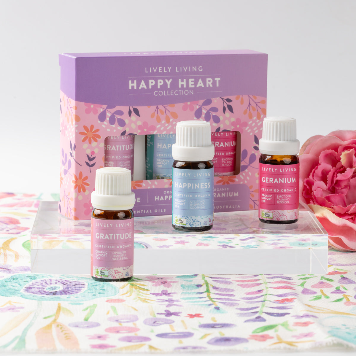 Happy Heart trio of Gratitude, Happiness and Geranium bottles on a clear stand with a pink rose