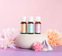 Lively Living trio of essential oils. Spearmint, Fresh Home, Calm and Unwind. Sitting on a white rock and pink background surrounded by pink and orange flowers. 