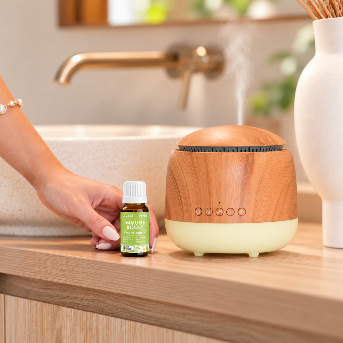 Hand placing a bottle of Lively Living Immune Boost organic essential oil beside a round wood-look diffuser releasing mist on a timber bathroom vanity with a stone basin and brass tap