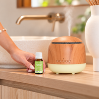 Hand placing a bottle of Lively Living Immune Boost organic essential oil beside a round wood-look diffuser releasing mist on a timber bathroom vanity with a stone basin and brass tap