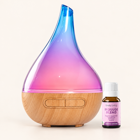 A teardrop-shaped essential oil diffuser with a wood-grain base and a translucent top that gradients from blue to pink to purple, shown next to a small bottle of Lively Living Indulge Blend essential oil on a light beige background.