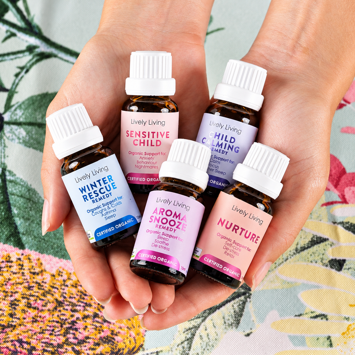 Two hands holding five Lively Living certified organic oils, Winter Rescue, Sensitive Child, Child Calming, Aroma Snooze and Nurture, over a botanical print fabric